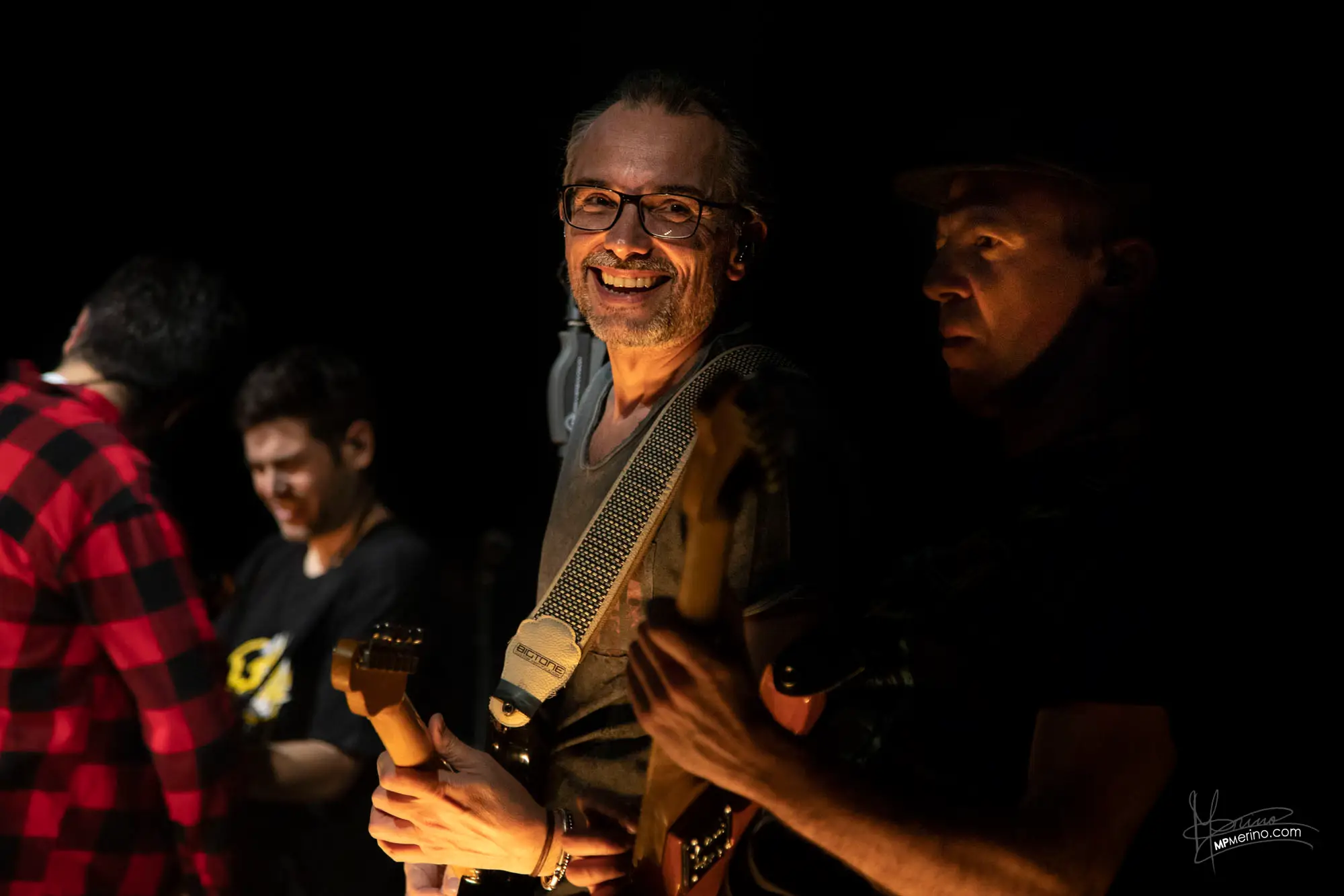 Concert photograph, members of the band Estopa play while one of them looks and smiles at the camera