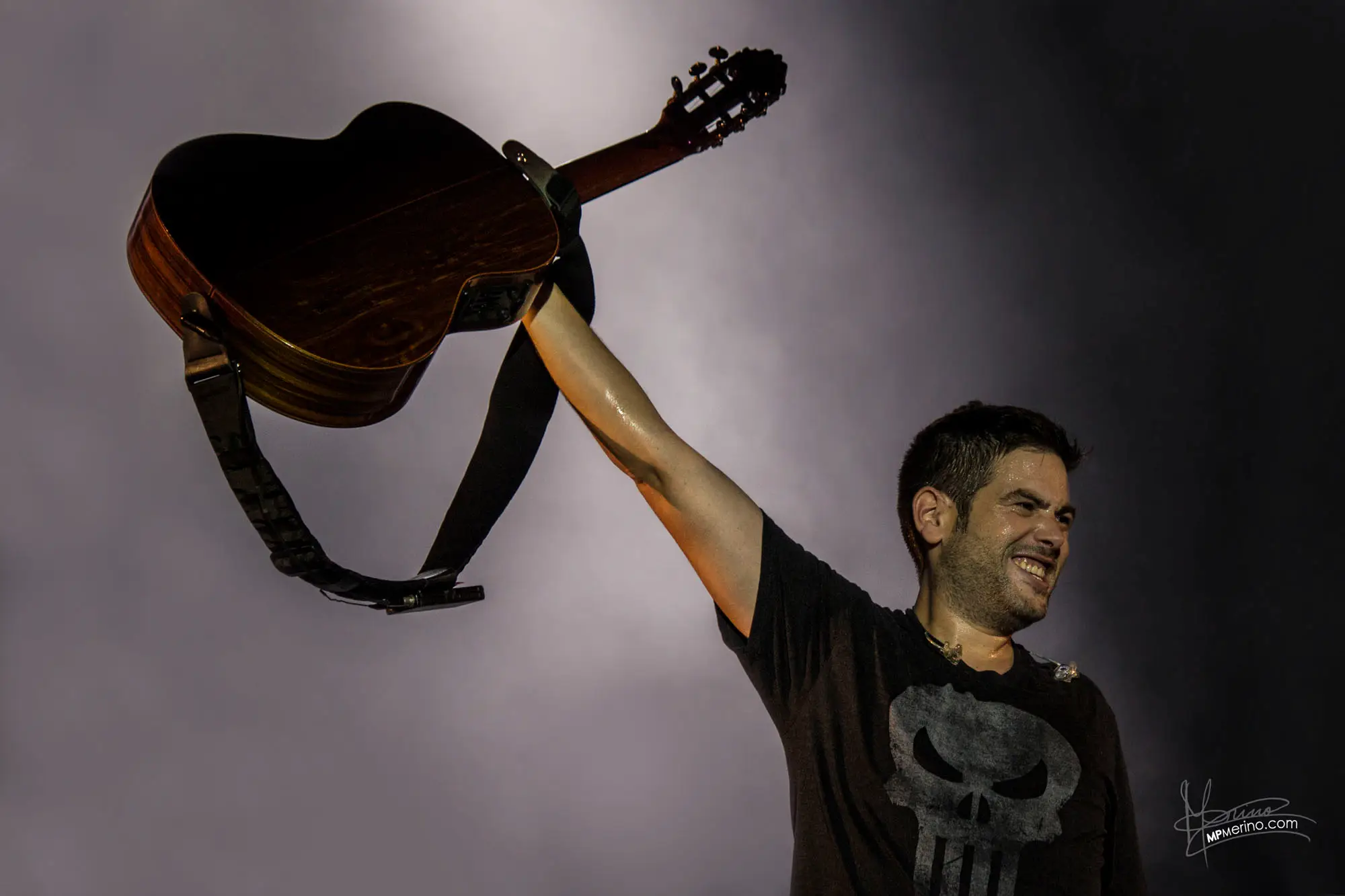 Concert photograph, a member of Estopa raises his guitar and smiles looking at the audience