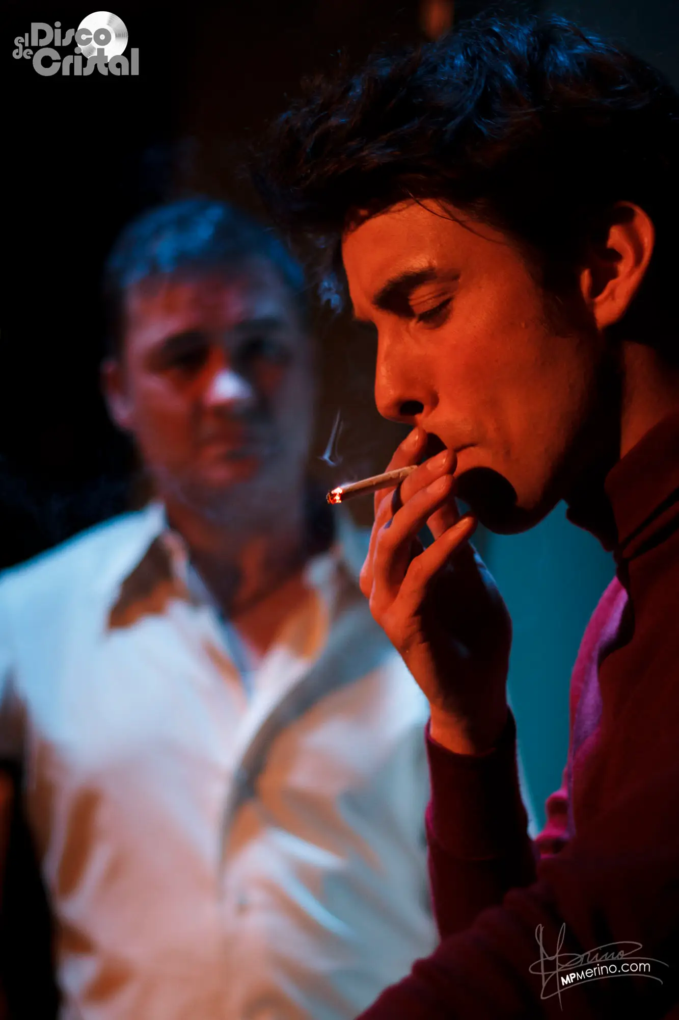 Theatre photograph, an actor smokes a cigarette while another actor watches him with a serious look on his face